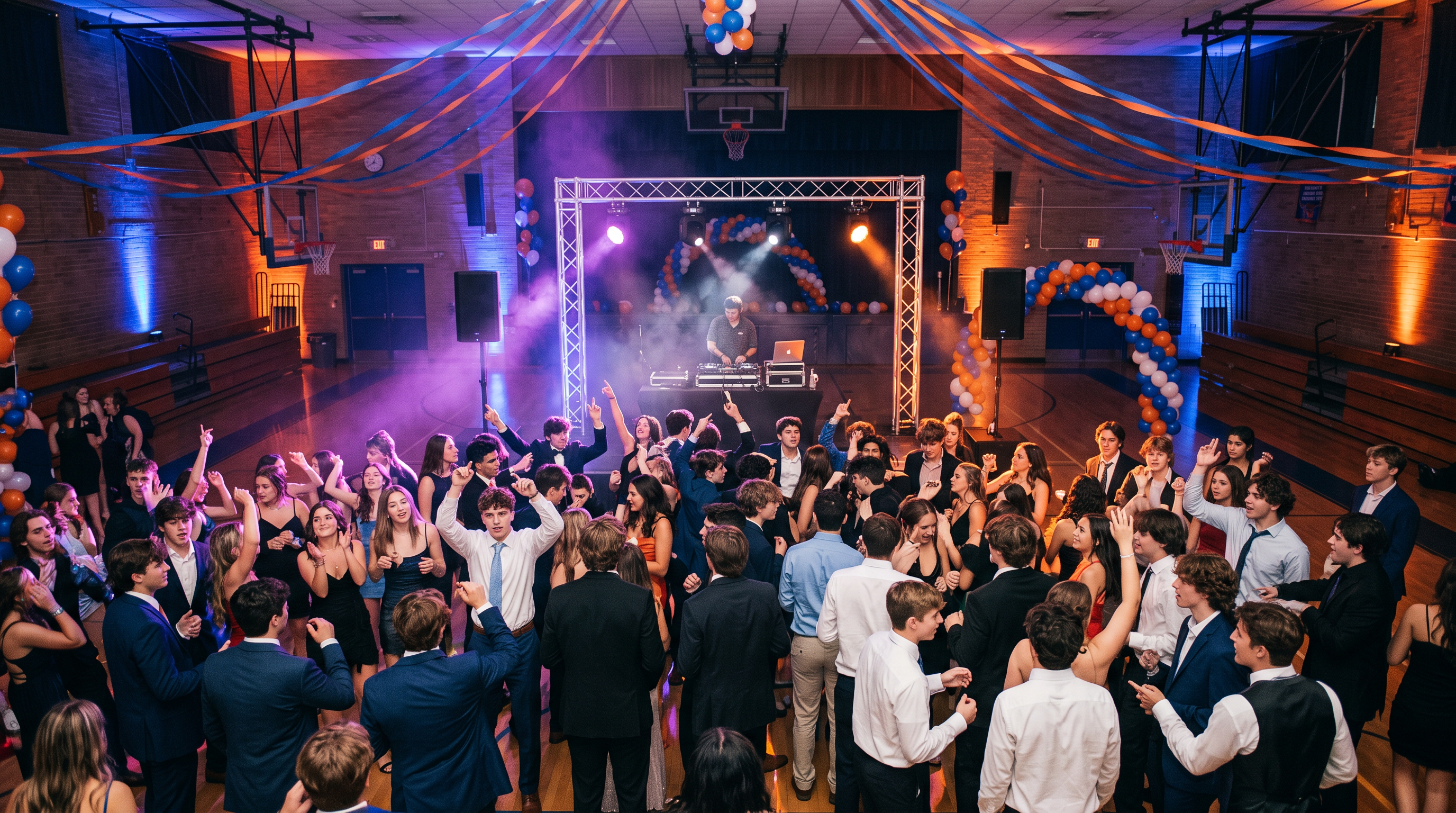 DJ setup with speakers and colorful uplighting at a high school homecoming dance in a decorated gymnasium with students dancing under blue and orange lights